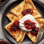 Thin golden Swedish pancakes folded into quarters, topped with whipped cream and lingonberry jam on a black plate with fork.