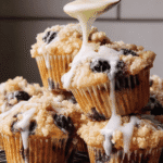 Stack of lemon blueberry muffins with streusel topping being drizzled with fresh lemon glaze from a spoon
