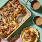 Cinnamon Roll Casserole with a serving spoon and a plated slice topped with icing and pecans, next to two cups of coffee.