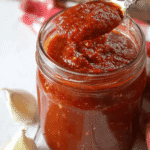Thick homemade pizza sauce in a glass jar with spoon, surrounded by fresh garlic cloves on a rustic wooden table.