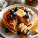Stack of fluffy buttermilk pancakes topped with butter, maple syrup, and blueberries, with a fork holding a bite-sized piece on a white plate