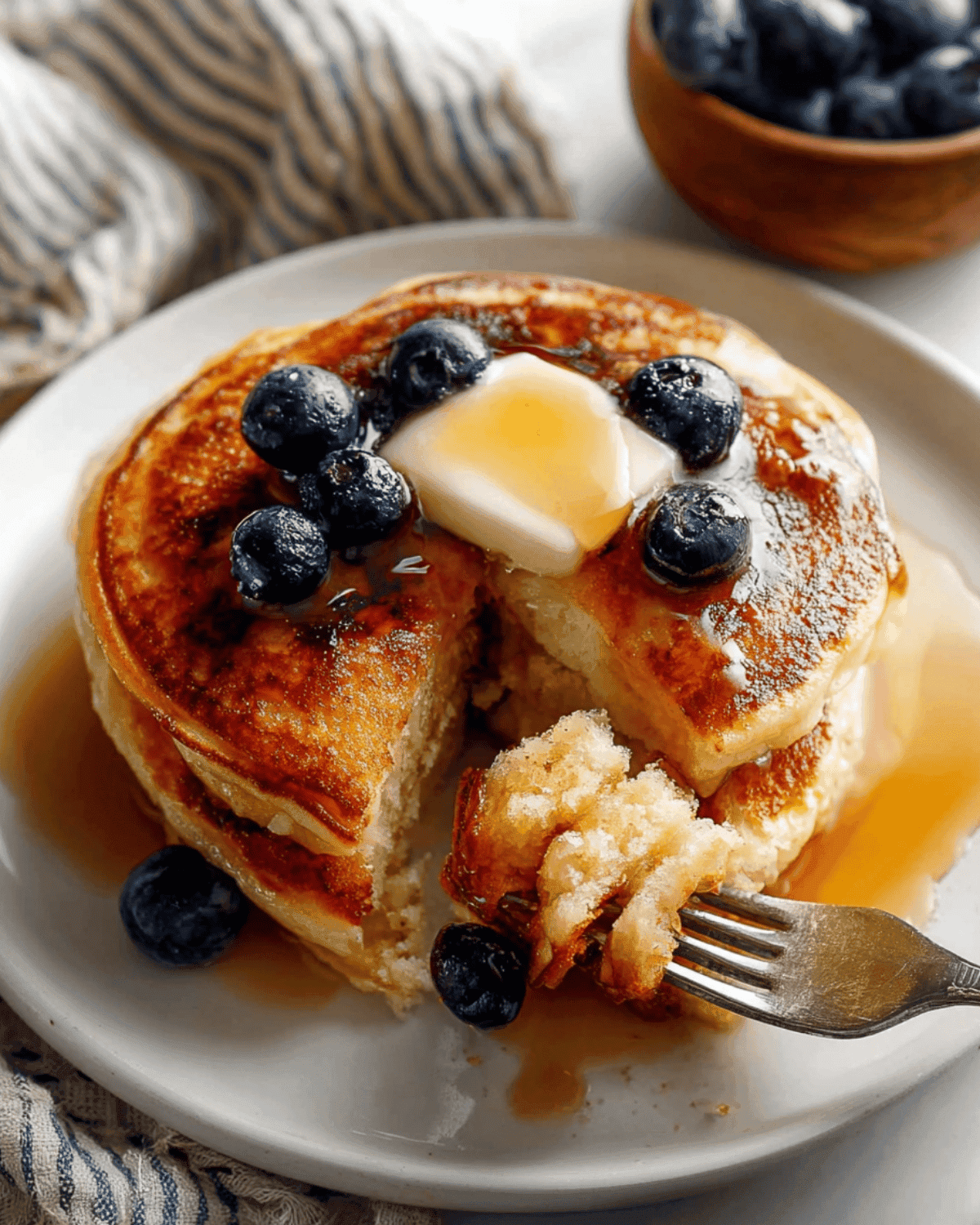 Fluffy Buttermilk Pancakes with Blueberries and Maple Syrup Stack of fluffy buttermilk pancakes topped with butter, maple syrup, and blueberries, with a fork holding a bite-sized piece on a white plate