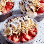 Three Copycat Acai Playa Bowls in clear containers, topped with sliced bananas, fresh strawberries, coconut flakes, granola, and chocolate drizzle.