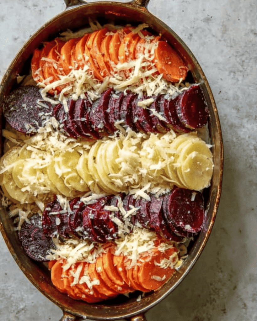 Unbaked root vegetable gratin with rows of sweet potatoes, beets, and Yukon gold potatoes topped with shredded cheese.