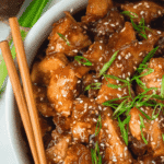 Close-up of sticky ginger chicken with sesame seeds and green onions served in a white bowl with chopsticks.