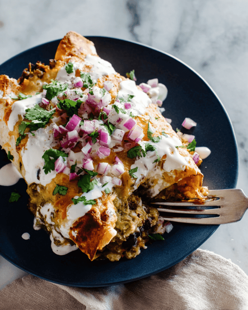 Close-up of Black Bean Sweet Potato Enchilada topped with sour cream, cilantro, and red onion on a dark plate