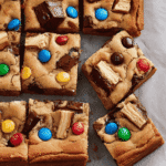 Fresh blondie squares topped with M&M’s and chocolate chunks, placed on parchment paper.