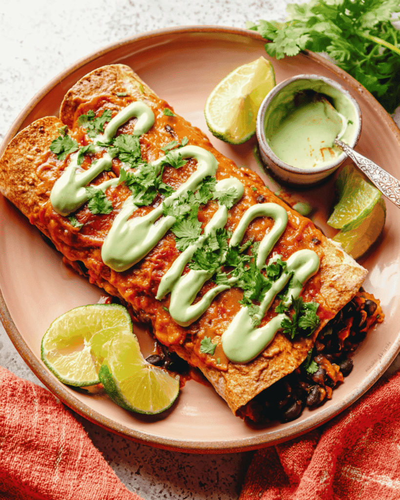 Plated pumpkin black bean enchiladas garnished with avocado crema, cilantro, and lime wedges