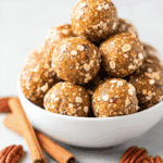 White bowl stacked with pumpkin oat energy bites surrounded by pecans and cinnamon sticks.