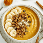 Pumpkin smoothie bowl topped with banana slices, granola, pumpkin seeds, and coconut flakes in a speckled ceramic bowl