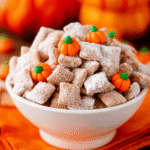 Bowl of pumpkin spice puppy chow with candy pumpkins on an orange cloth for a festive holiday snack mix