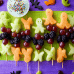 Spooky Halloween Fruit Kabobs with ghost melon, bat cantaloupe, grapes, and blackberries on a colorful mat.