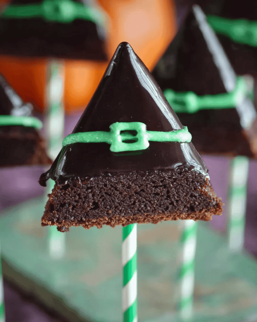 Halloween brownie pop shaped like a witch hat with shiny black icing and green icing buckle on a striped stick.