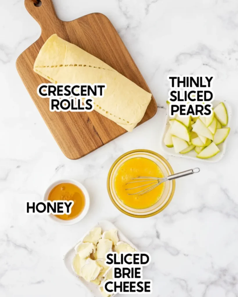 Flat lay of ingredients for Brie Pear Bites including crescent roll dough, thinly sliced pears, brie cheese, beaten egg, and honey on a marble surface.