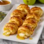 Golden baked Brie Pear Crescent Bites drizzled with honey on a white serving plate, with green pears and holiday decor in the background.