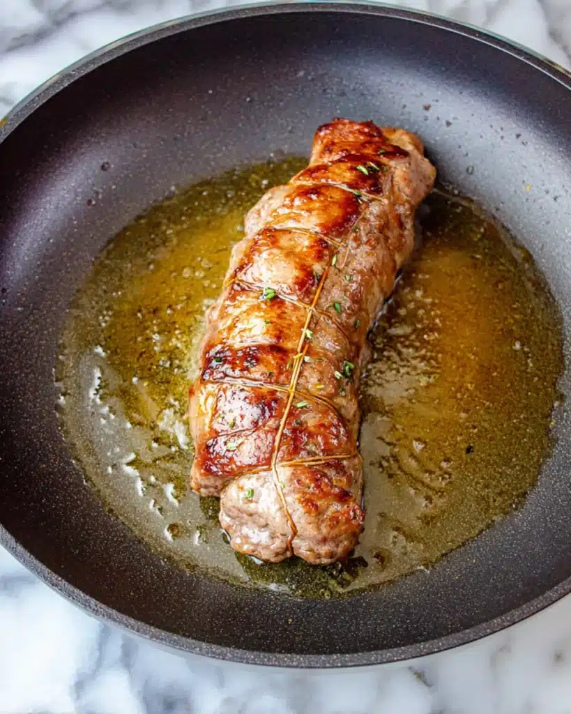 Rolled beef braciole seared to golden brown in olive oil inside a skillet.