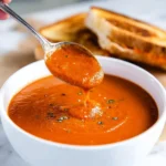Close-up of a spoon lifting thick, creamy tomato soup from a white bowl with grilled cheese in the background.