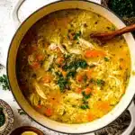 Top-down view of a white pot filled with chicken and rice soup with wooden spoon and herbs