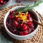 Homemade cranberry sauce in a white bowl with orange zest and rosemary garnish