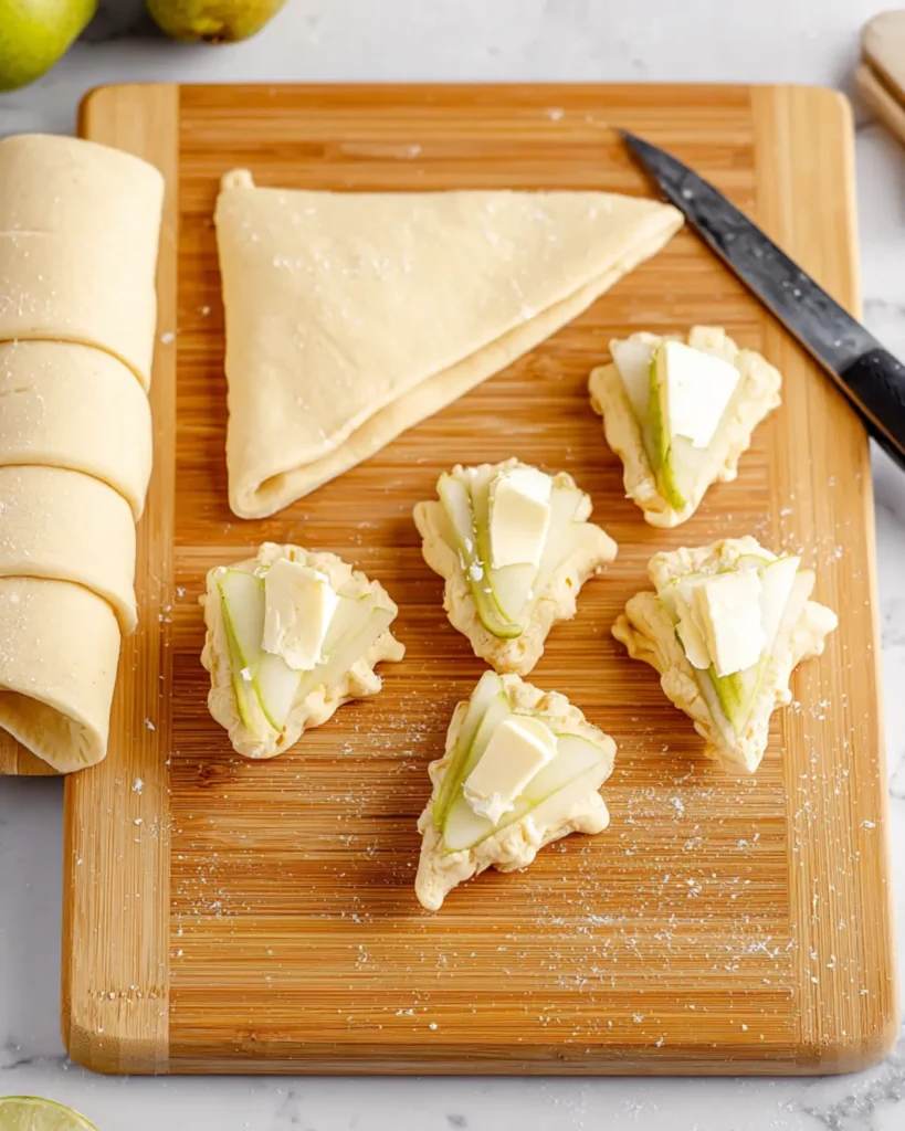 Preparing crescent dough triangles topped with thin pear slices and soft cheese on a wooden cutting board, ready to roll into pastries.