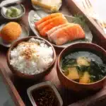 Japanese meal tray with miso soup, steamed rice, salmon sashimi, tamagoyaki, and dipping sauce arranged neatly on wooden plates