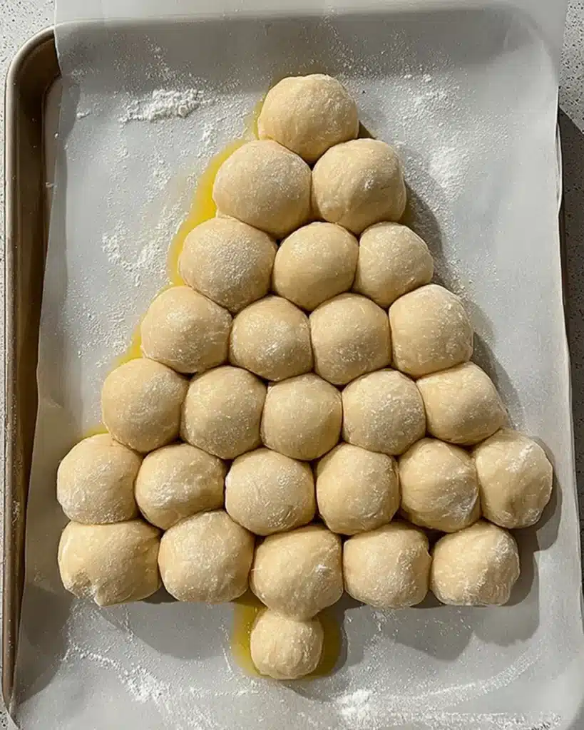 Raw pizza dough balls arranged in a Christmas tree shape on parchment paper for Cheesy Christmas Tree Bread.