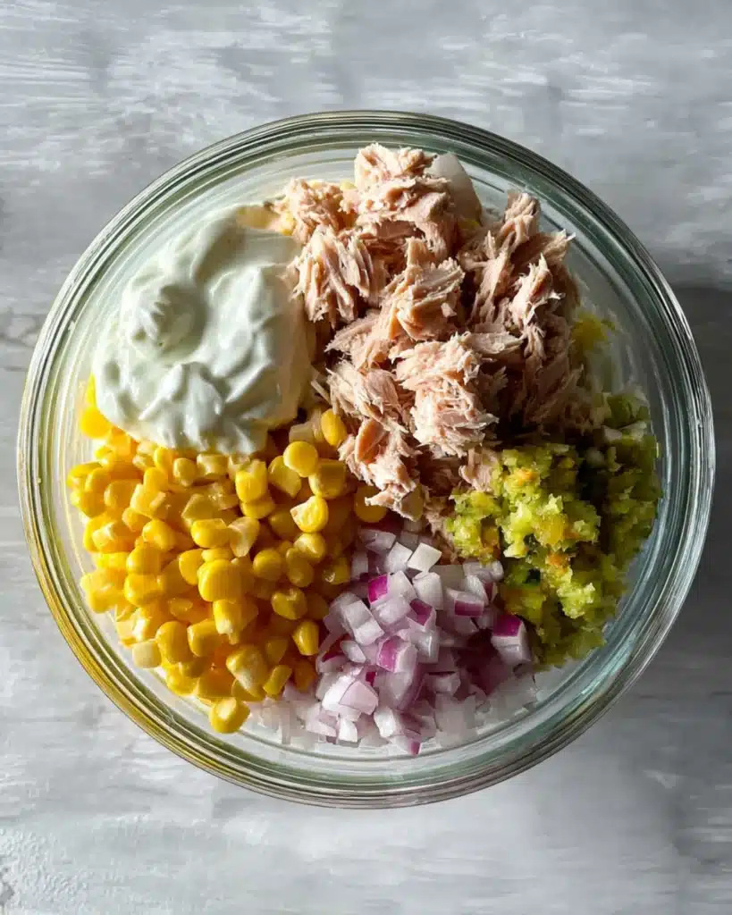 Top-down view of tuna melt wrap ingredients in a glass bowl including tuna, corn, yogurt, relish, and onions.