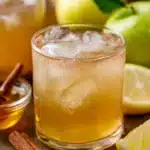 Glass of apple cider vinegar drink with ice, lemon slices, and cinnamon sticks on a wooden table.