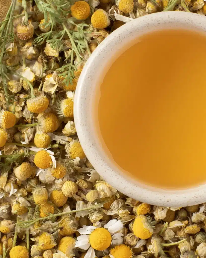 Overhead view of dried chamomile flowers with a white cup of brewed golden tea.