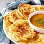 Flaky roti canai served with a bowl of creamy curry on a blue striped cloth.