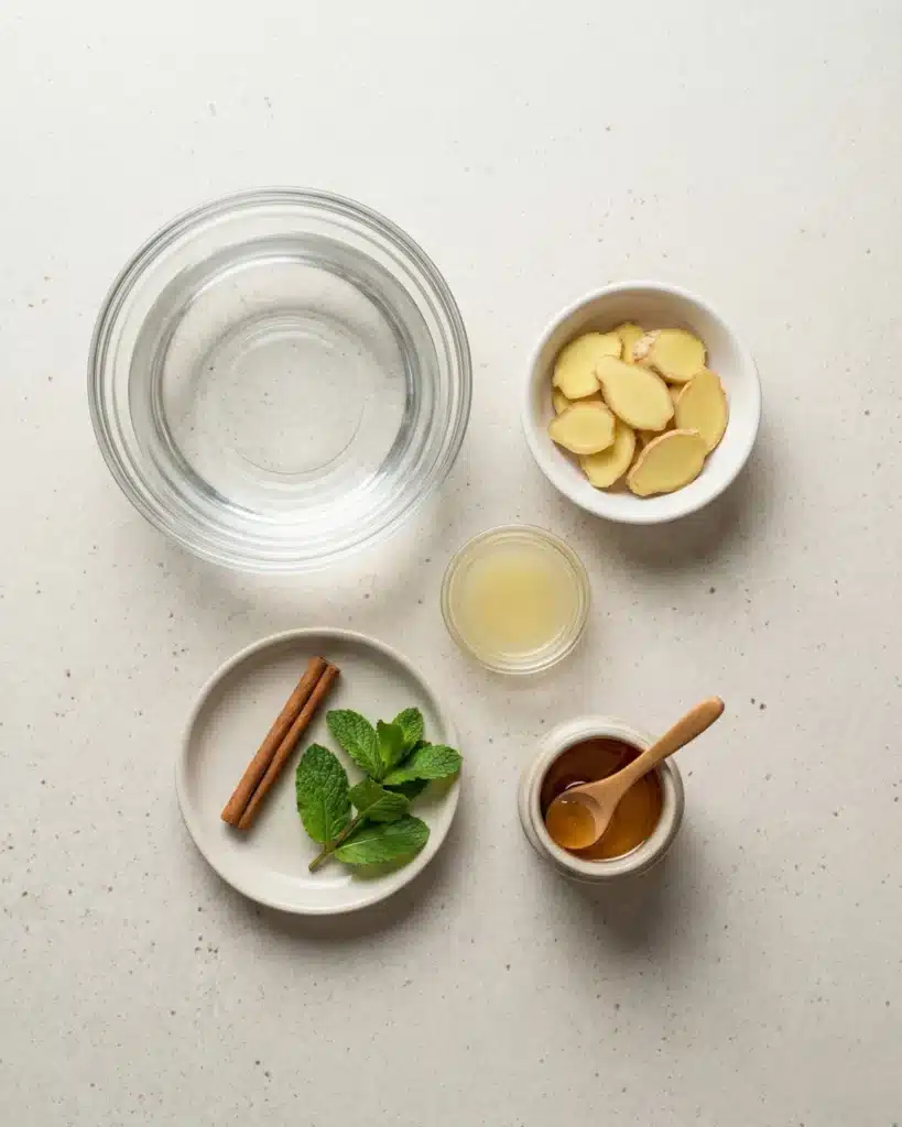Top view of fresh ginger slices, mint leaves, cinnamon stick, lemon juice, honey, and a bowl of water arranged on a white countertop for making ginger tea.