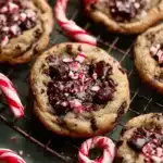 Close-up of freshly baked peppermint chocolate chip cookies topped with melted chocolate and crushed candy canes cooling on a wire rack.