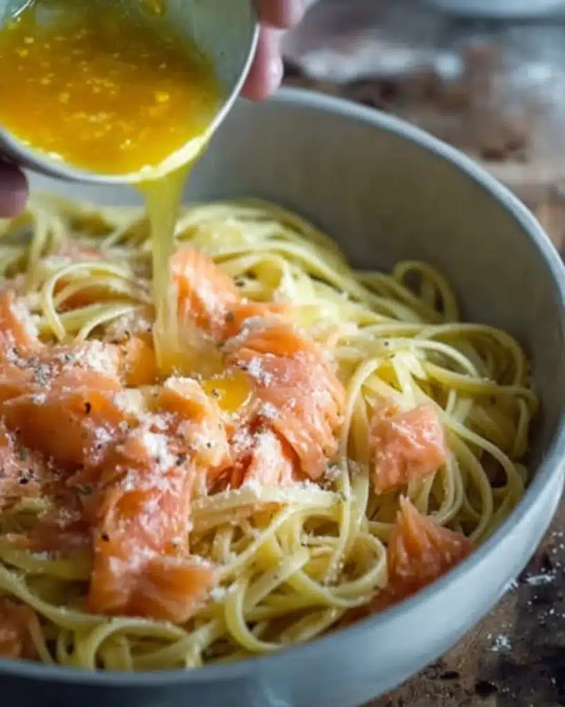 Hand pouring egg and Parmesan sauce over linguine pasta with smoked salmon for carbonara preparation.