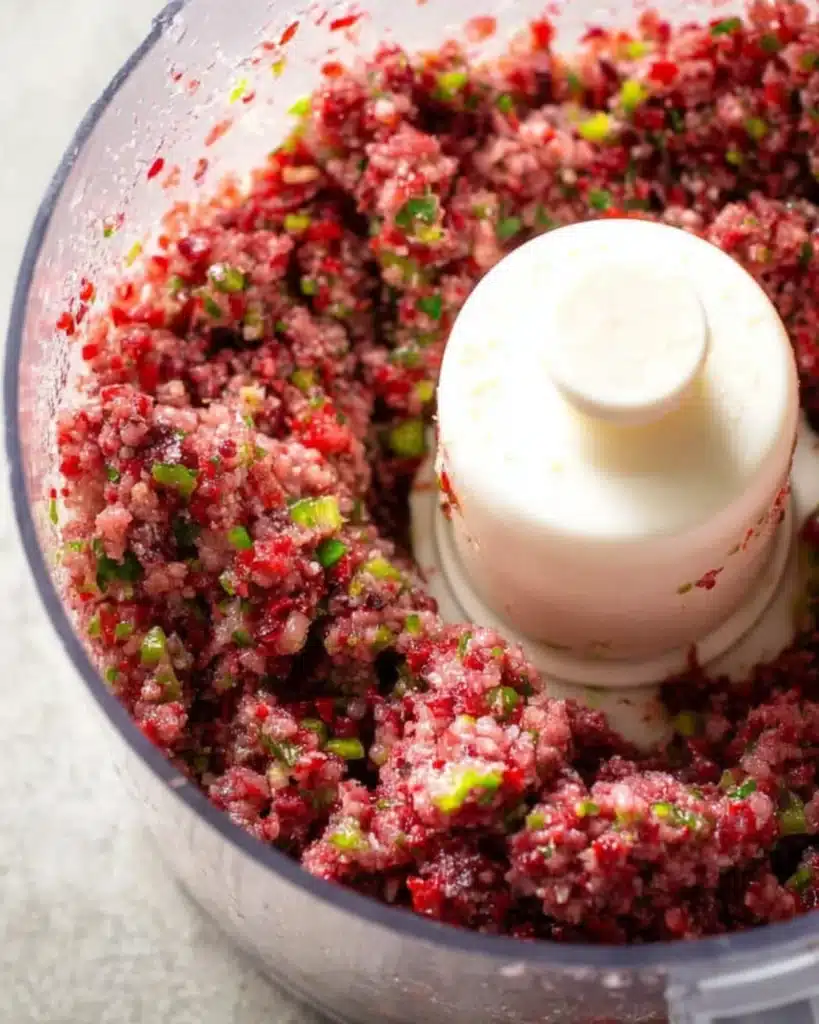 Pulsed Cranberry Salsa in a food processor showing ruby cranberries with green jalapeño and herb pieces