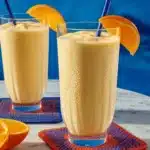 Close-up of two tall glasses filled with thick, creamy Orange-Mango Smoothie topped with orange slice garnish and blue straws.