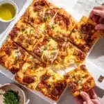 Overhead view of square pizza slices topped with golden cheese, herbs, and olive oil drizzle