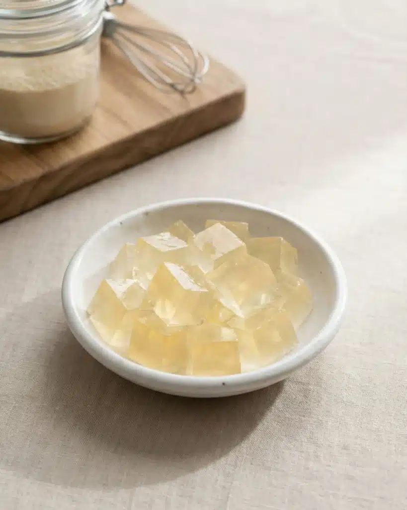 Clear gelatin cubes in a white ceramic bowl beside a jar of powdered gelatin on a wooden board