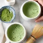 Two creamy cups of matcha latte with frothy green tops placed on ceramic saucers beside a small bowl of powdered tea