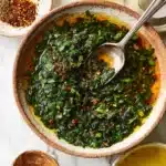 A rustic bowl of chimichurri sauce being stirred with a spoon, highlighting fresh parsley and olive oil texture.