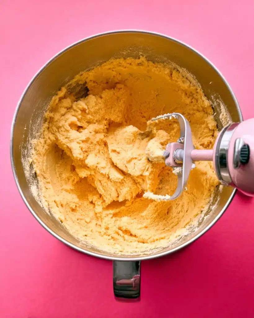 Yellow sugar cookie dough being mixed in a metal bowl with a pink mixer paddle.