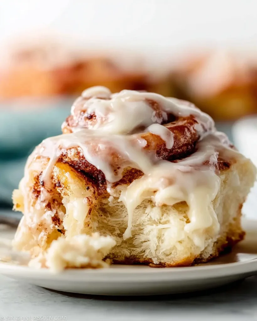 Soft pull apart cinnamon bun on a white plate with glossy icing dripping over the top