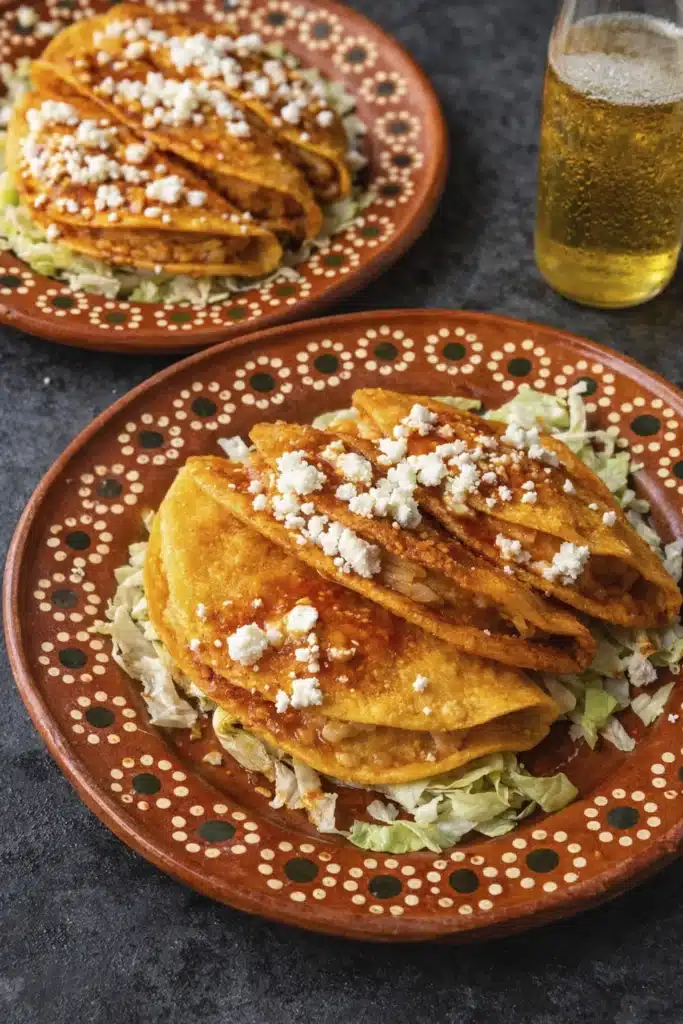 Authentic cheesy potato tacos served on a plate with lettuce, queso fresco, and a refreshing drink