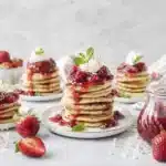 Coconut ricotta pancakes served on multiple plates with strawberry syrup and fresh berries