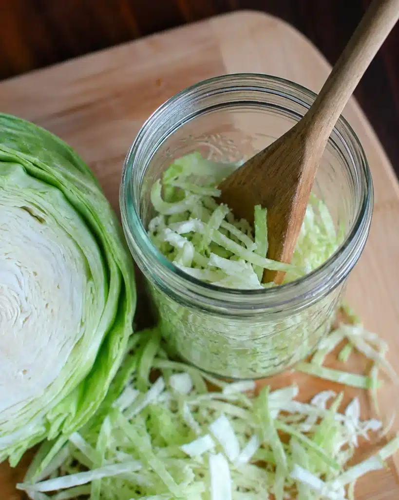 fresh cabbage cut in half next to a jar filled with shredded pieces and a wooden spoon
