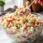 Spoon lifting a bite of creamy pasta salad with chickpeas and vegetables from a glass bowl