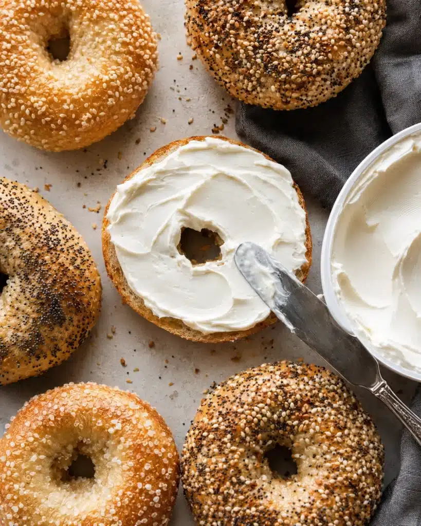 Sliced bagel topped with smooth cream cheese being spread with a knife on a light surface