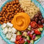 Close up of hummus, quinoa, chickpeas, feta, olives, and fresh vegetable salad in a patterned bowl