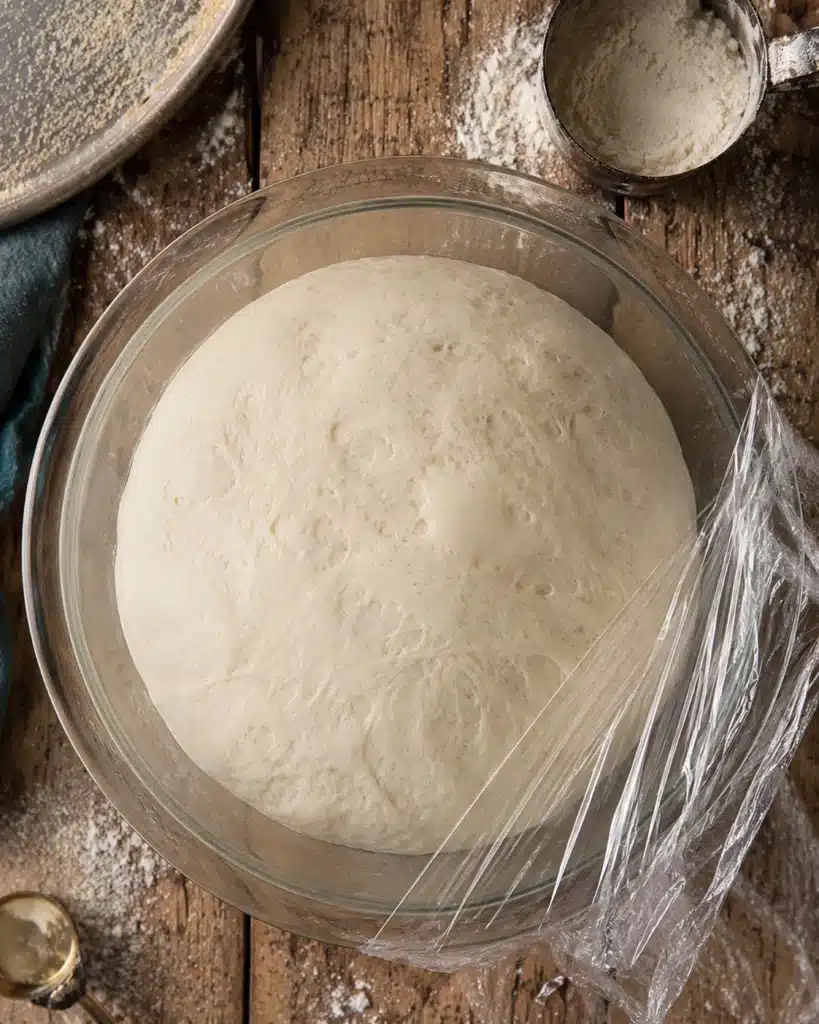 Dough rising in a glass bowl covered with plastic wrap on a wooden table