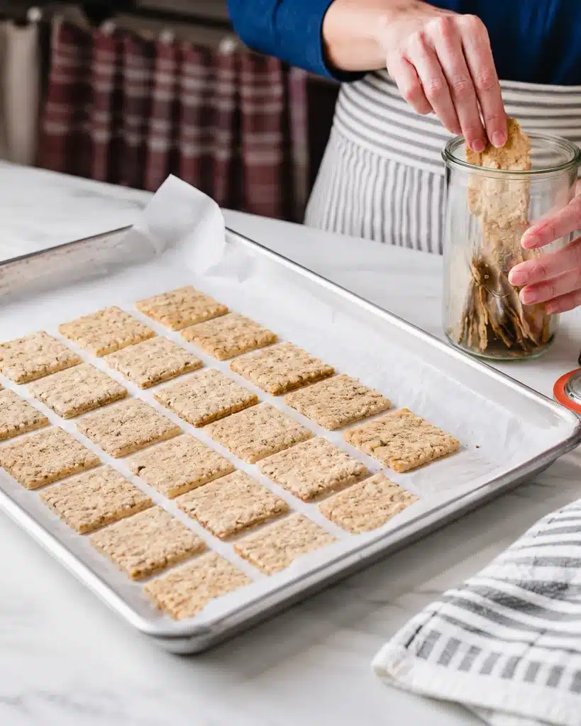 hand placing baked crackers into a glass jar beside a tray of freshly baked crackers