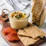 creamy hummus served in a glass jar with crackers and sliced cured meat on a wooden board
